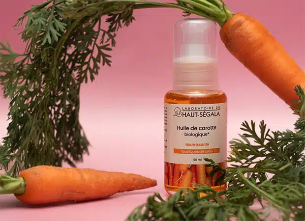 Flacon d'Huile de Carotte biologique 50ml du Laboratoire Haut-Ségala, entouré de carottes fraîches et de leurs fanes.