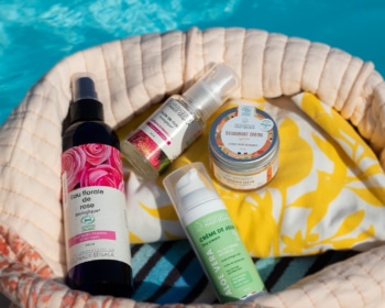Un sac de plage en tissu beige et fleuri contient des flacons de cosmétiques naturels été de la marque Haut-Ségala, posé au bord d'une piscine. On distingue clairement une Eau florale de rose biologique, une Crème de Jour Aloe Vera, un Déodorant Crème et une Huile de ricin. L'image illustre une routine beauté estivale avec des produits de beauté naturels piscine de la gamme bio Haut-Ségala.