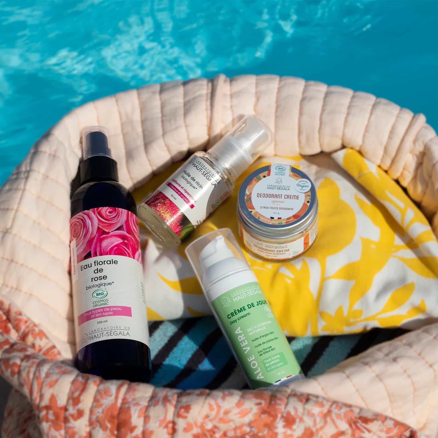 Un sac de plage en tissu beige et fleuri contient des flacons de cosmétiques naturels été de la marque Haut-Ségala, posé au bord d'une piscine. On distingue clairement une Eau florale de rose biologique, une Crème de Jour Aloe Vera, un Déodorant Crème et une Huile de ricin. L'image illustre une routine beauté estivale avec des produits de beauté naturels piscine de la gamme bio Haut-Ségala.