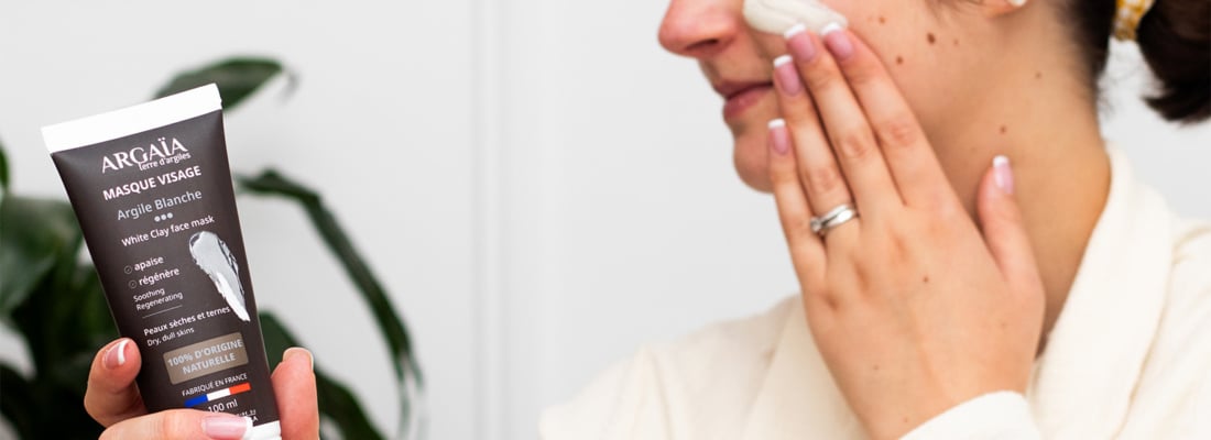 Une personne applique le Masque Visage Argile Blanche Argaïa sur son visage tout en tenant le tube du produit. L'étiquette est visible et indique "Masque Visage Argile Blanche", "purifie", "matifie", "White Clay face mask", "Peaux mixtes à grasses", "100% D'ORIGINE NATURELLE", et "FABRIQUÉ EN FRANCE". L'image représente un masque argile blanche peaux grasses et un masque visage purifiant naturel.