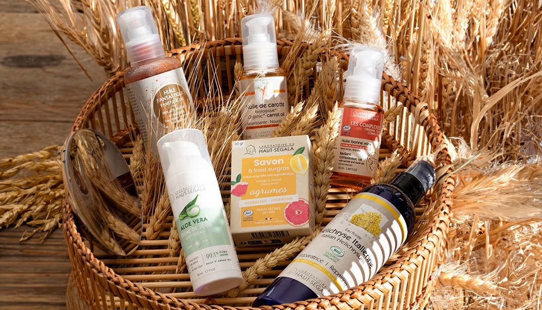 Panier en osier contenant plusieurs produits cosmétiques du Laboratoire Haut-Ségala (Gel Aloe Vera, savon, huiles, eau florale). Arrière-plan champêtre avec des épis de blé.