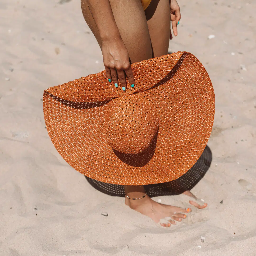 Femme tenant un grand chapeau de plage orange sur le sable qui part pour une séance de bronzage.