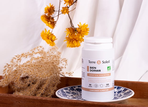 Flacon de complément alimentaire "BIEN DORMIR" de Terre et Soleil sur une soucoupe, entouré de fleurs séchées, pour illustrer une solution naturelle pour le sommeil. L'étiquette indique "120 gélules" pour "30 jours" et le logo AB.