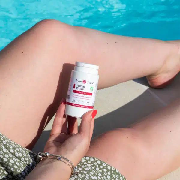 Une main féminine tenant une bouteille blanche de Complément Alimentaire Ginkgo Biloba Terre & Soleil au bord d'une piscine. L'étiquette est visible, indiquant "Ginkgo Biloba" et "Complément Alimentaire". L'image évoque le soutien cognitif naturel et le bien-être estival apportés par le ginkgo biloba mémoire concentration.