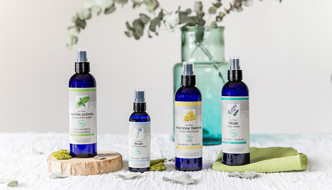 Quatre flacons d'eaux florales du Laboratoire Haut-Ségala posés sur une table. De gauche à droite, les étiquettes sont "Menthe poivrée", "Bleuet", "Hélichryse italienne" et "Sauge". L'image illustre une collection de soins naturels pour le visage.