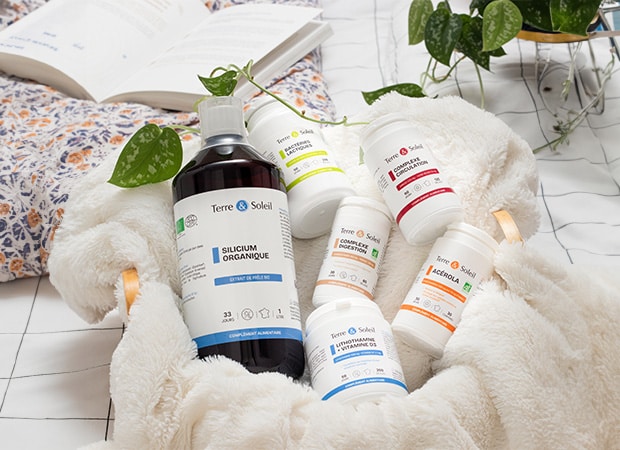 Une composition de six flacons de compléments alimentaires Terre & Soleil posés sur une couverture blanche duveteuse, avec un livre et une plante en arrière-plan. Les produits visibles incluent "Silicium Organique", "Bactéries Lactiques", "Complexe Circulation", "Complexe Digestion", "Lithothamne + Vitamine D3", et "Acérola". L'image représente une gamme santé intérieure naturelle de compléments alimentaires bien-être pour une vitalité quotidienne.