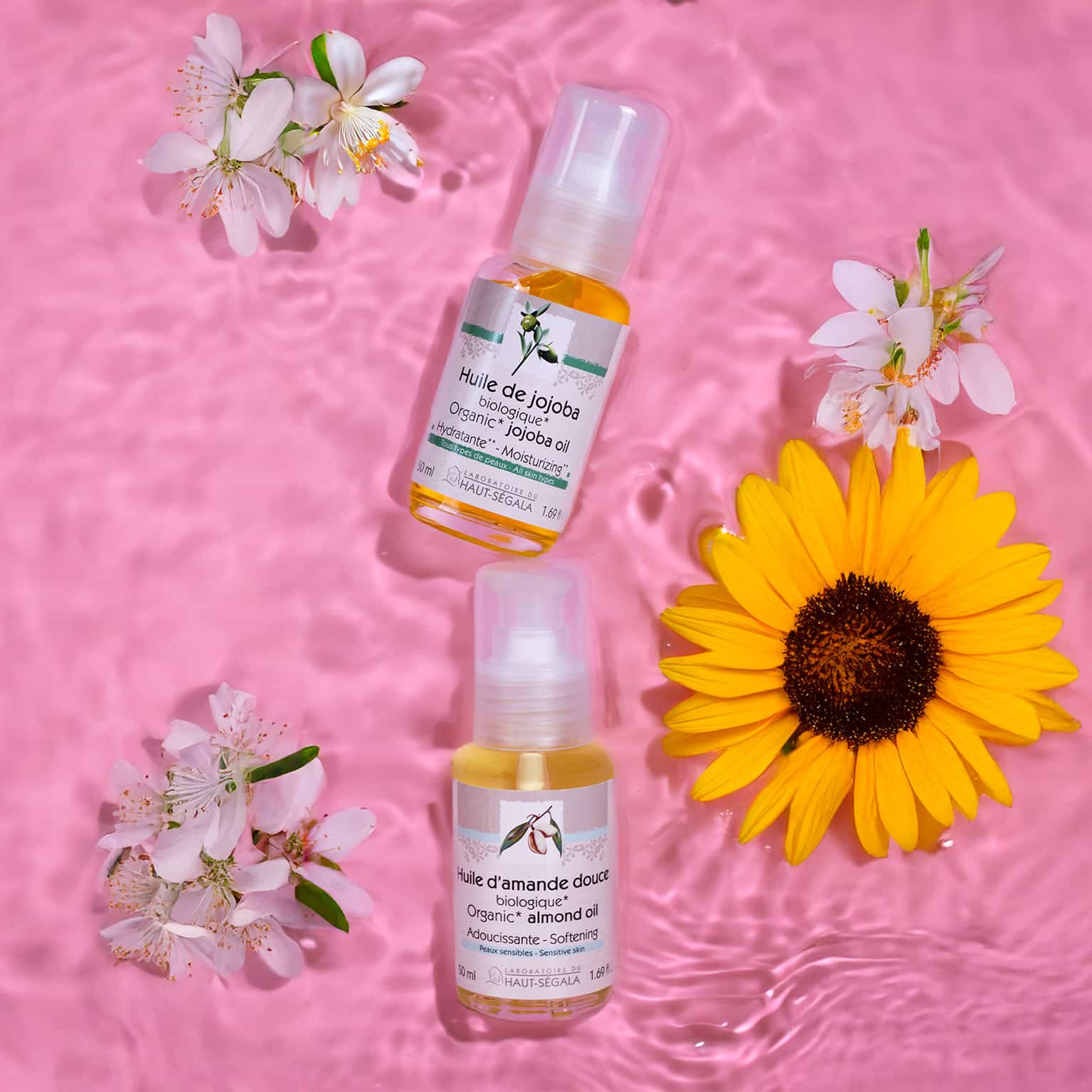 Deux flacons d'huiles végétales biologiques Haut-Ségala : Huile de Jojoba et Huile d'Amande Douce, posés sur l'eau avec des fleurs.