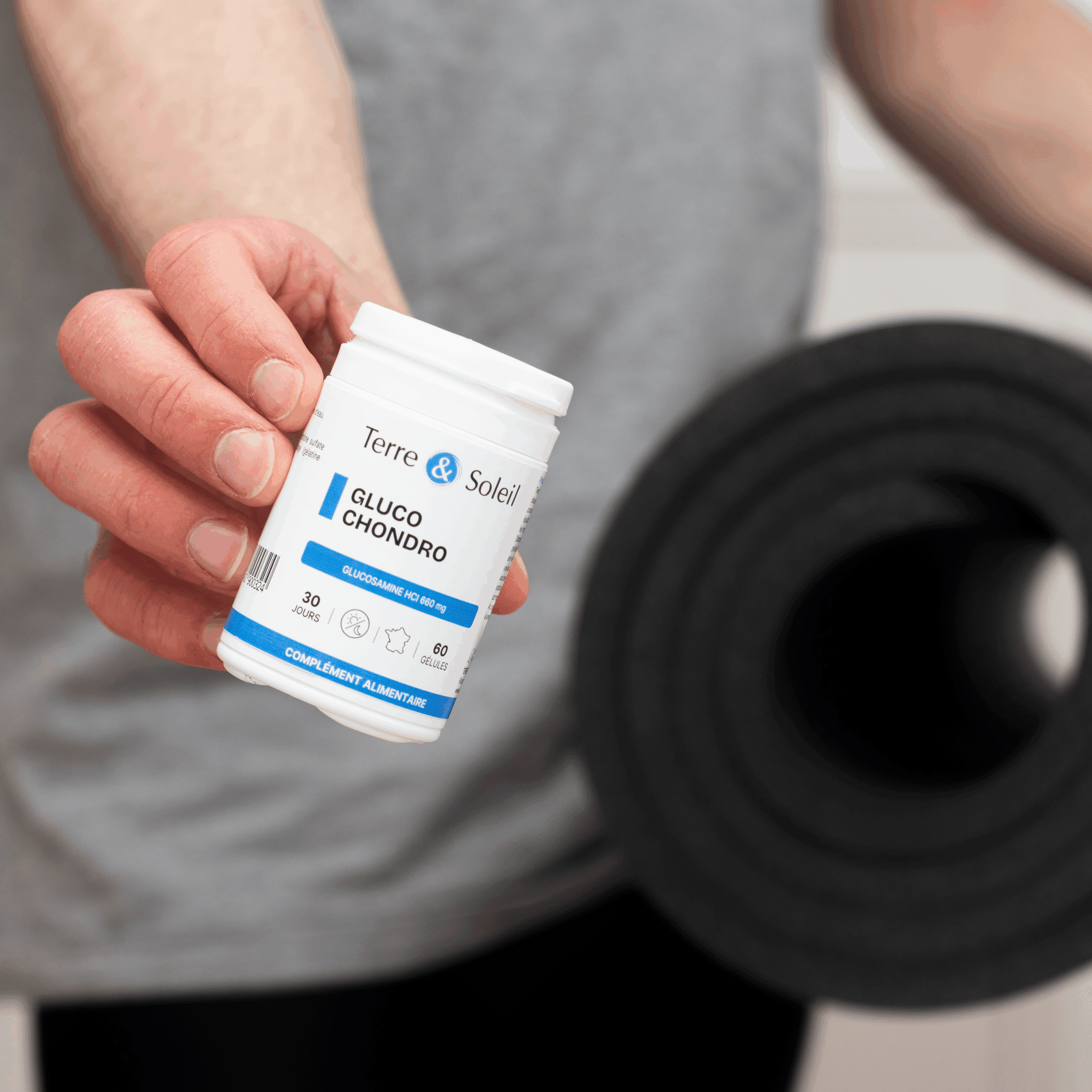 Personne tenant un flacon de complément alimentaire "Gluco Chondro" de Terre et Soleil, avec un tapis de sport enroulé sous le bras, pour illustrer le soutien des articulations après l'activité physique.