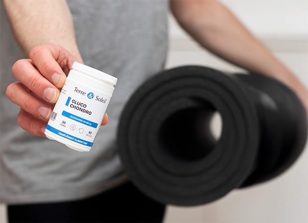 Personne tenant un flacon de complément alimentaire "Gluco Chondro" de Terre et Soleil, avec un tapis de sport enroulé sous le bras, pour illustrer le soutien des articulations après l'activité physique.