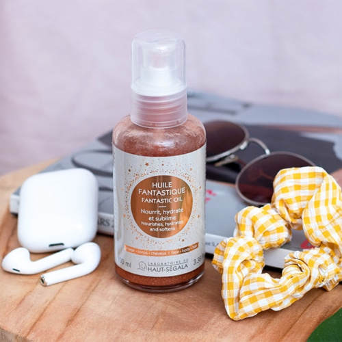 Une bouteille de "LABORATOIRE DU HAUT-SÉGALA Huile Fantastique" est posée sur une table en bois. L'étiquette indique "Fantastic oil", "Nourrit, hydrate et sublime", "Nourishes, hydrates and softens", et "Visage - Corps - Cheveux", "100% naturel". Des lunettes de soleil, des écouteurs sans fil et un chouchou jaune à carreaux sont en arrière-plan. L'image représente une huile sèche multi-usage naturelle et un soin visage corps cheveux pour éclat peau cheveux.