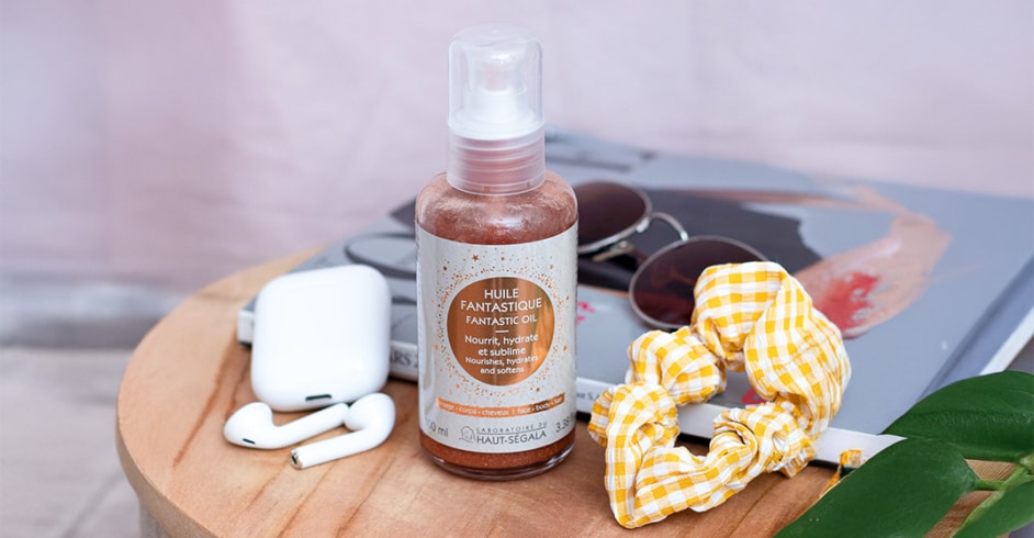 Une bouteille de "LABORATOIRE DU HAUT-SÉGALA Huile Fantastique" est posée sur une table en bois. L'étiquette indique "Fantastic oil", "Nourrit, hydrate et sublime", "Nourishes, hydrates and softens", et "Visage - Corps - Cheveux", "100% naturel". Des lunettes de soleil, des écouteurs sans fil et un chouchou jaune à carreaux sont en arrière-plan. L'image représente une huile sèche multi-usage naturelle et un soin visage corps cheveux pour éclat peau cheveux.