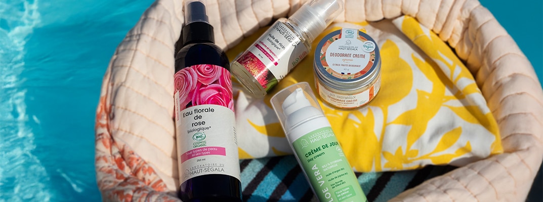 Un sac de plage en tissu beige et fleuri contient des flacons de cosmétiques naturels été de la marque Haut-Ségala, posé au bord d'une piscine. On distingue clairement une Eau florale de rose biologique, une Crème de Jour Aloe Vera, un Déodorant Crème et une Huile de ricin. L'image illustre une routine beauté estivale avec des produits de beauté naturels piscine de la gamme bio Haut-Ségala.