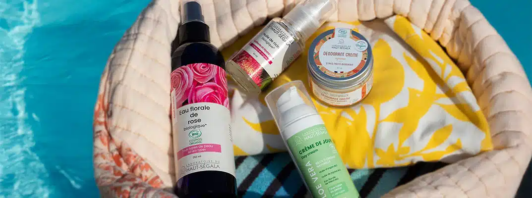 Un sac de plage en tissu beige et fleuri contient des flacons de cosmétiques naturels été de la marque Haut-Ségala, posé au bord d'une piscine. On distingue clairement une Eau florale de rose biologique, une Crème de Jour Aloe Vera, un Déodorant Crème et une Huile de ricin. L'image illustre une routine beauté estivale avec des produits de beauté naturels piscine de la gamme bio Haut-Ségala.