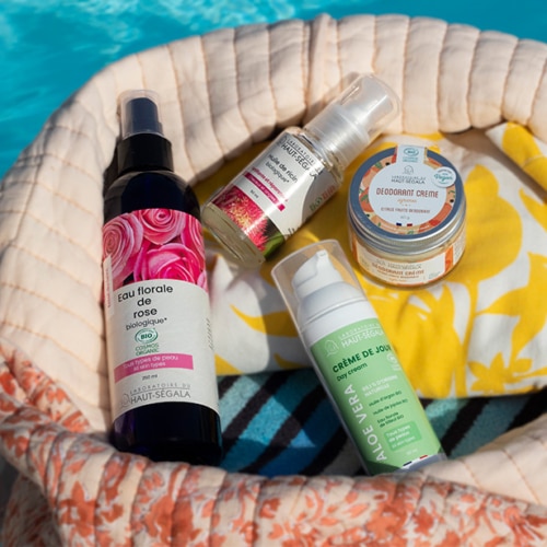 Un sac de plage en tissu beige et fleuri contient des flacons de cosmétiques naturels été de la marque Haut-Ségala, posé au bord d'une piscine. On distingue clairement une Eau florale de rose biologique, une Crème de Jour Aloe Vera, un Déodorant Crème et une Huile de ricin. L'image illustre une routine beauté estivale avec des produits de beauté naturels piscine de la gamme bio Haut-Ségala.