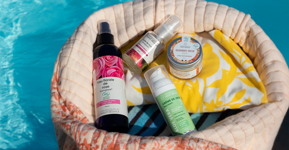 Un sac de plage en tissu beige et fleuri contient des flacons de cosmétiques naturels été de la marque Haut-Ségala, posé au bord d'une piscine. On distingue clairement une Eau florale de rose biologique, une Crème de Jour Aloe Vera, un Déodorant Crème et une Huile de ricin. L'image illustre une routine beauté estivale avec des produits de beauté naturels piscine de la gamme bio Haut-Ségala.