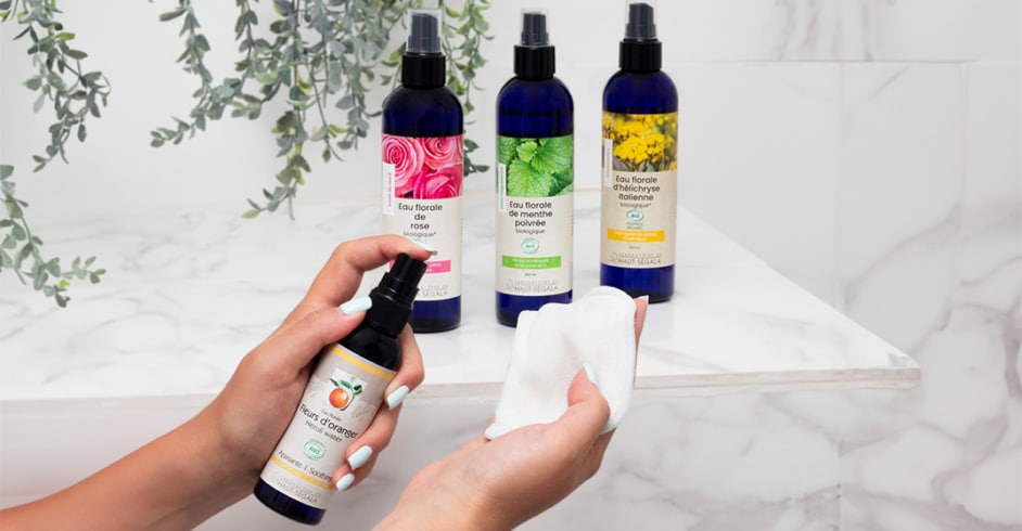 Personne vaporisant de l'eau florale de fleur d'oranger bio Laboratoire Haut-Ségala sur un coton, avec des bouteilles d'eau de rose, menthe poivrée et hélichryse en arrière-plan sur une étagère de salle de bain.