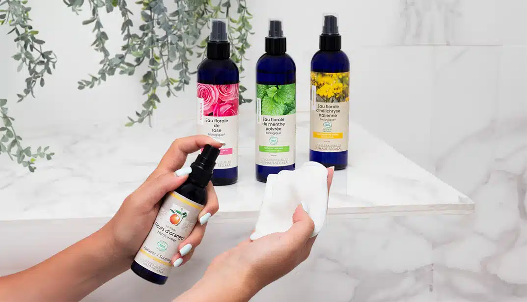 Personne vaporisant de l'eau florale de fleur d'oranger bio Laboratoire Haut-Ségala sur un coton, avec des bouteilles d'eau de rose, menthe poivrée et hélichryse en arrière-plan sur une étagère de salle de bain.